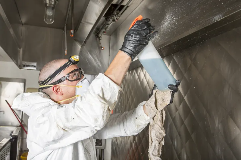 Technician in white coveralls, goggles and a headlamp reaching up with a spray bottle and cloth to wipe the underside of a stainless steel exhaust canopy, seen from below