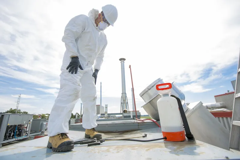 Technician in white coveralls, hard hat and respirator standing on a flat commercial roof beside an upblast exhaust fan and a pump sprayer under a cloudy sky