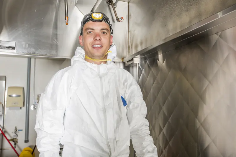 Hood cleaning technician in protective coveralls and a respirator standing in a commercial kitchen beside the cook line