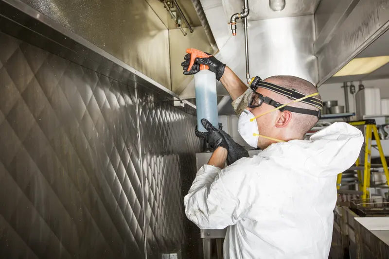 Technician spraying degreaser on the underside of a stainless steel commercial kitchen exhaust canopy with the baffle filters removed
