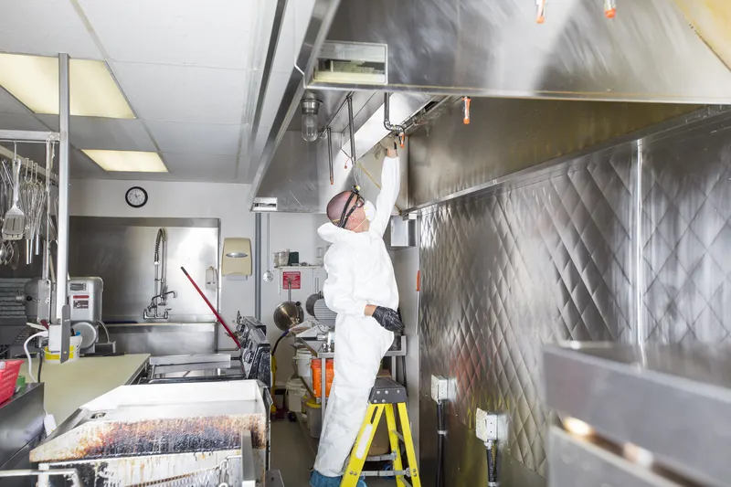 Hood cleaning technician on a step ladder cleaning a stainless steel canopy in a full commercial kitchen