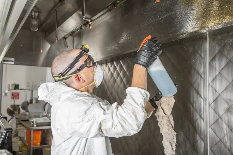 Technician in white coveralls, safety goggles and a headlamp wiping the underside of a stainless steel canopy hood with a cloth while holding a spray bottle in a commercial kitchen